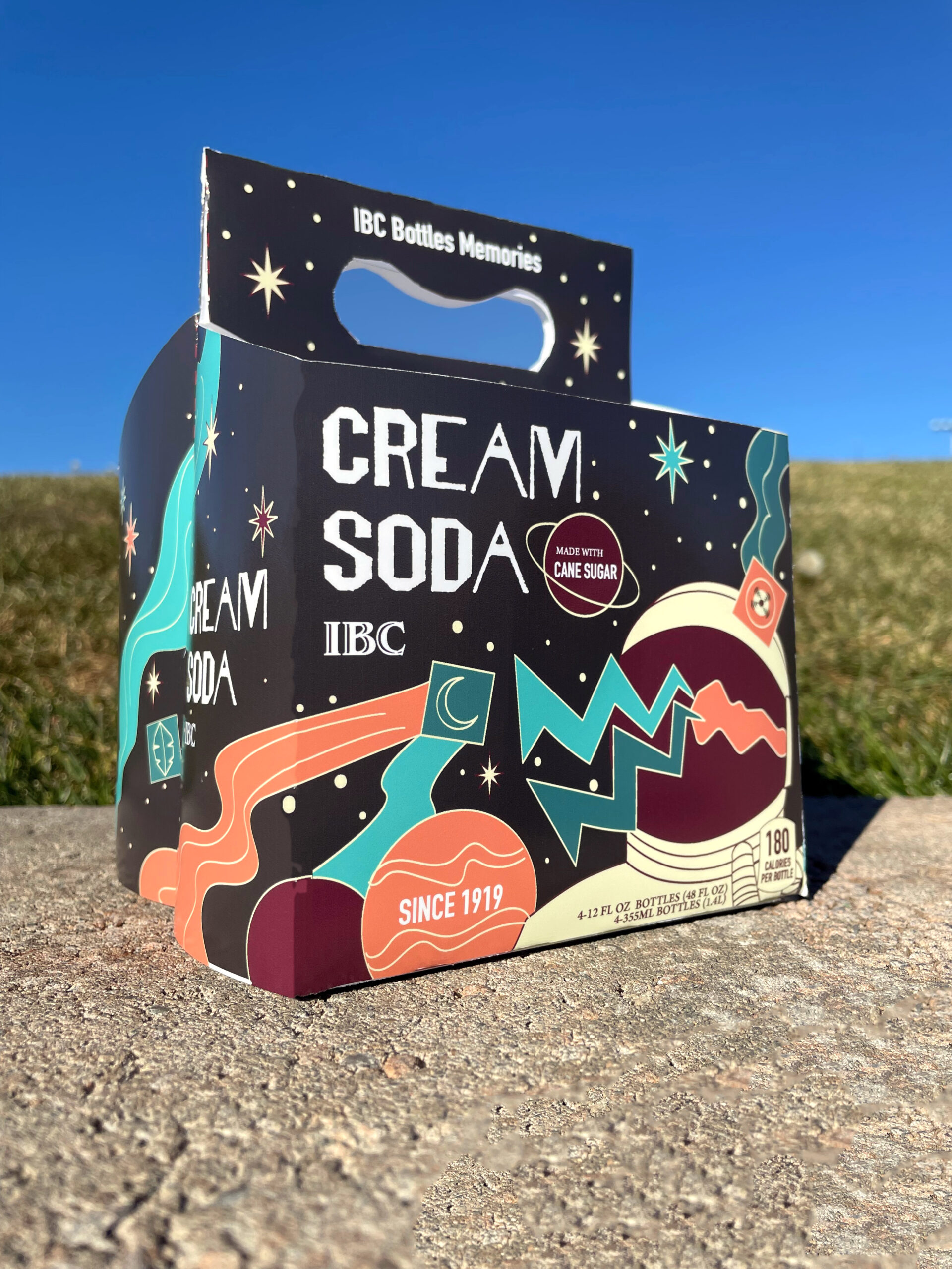 The four pack box sitting on cement; grass and sky are visible in the background. The four pack is black with illustration of space in bright oranges, reds, and blues. There is an astronaut, planets and stars. The copy reads Cream Soda.